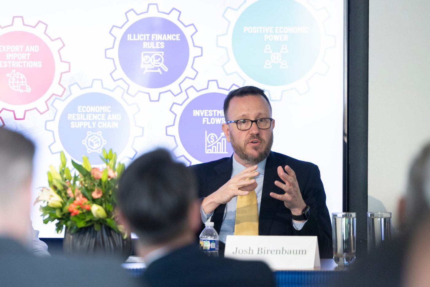 Josh Birenbaum speaking on a panel in front of a slide with gears labeled illicit finance rules, economic resilience and supply chain, investment flows, and positive economic power.